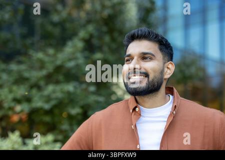 Ein lächelnder Indianer mit Bart, gekleidet in einem lässigen orangen Hemd, sieht aus der Kamera mit grünem Laub im Hintergrund. Stockfoto
