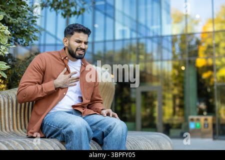 Ein Mann, der Schmerzen in der Brust hat, im Freien sitzt, wahrscheinlich ein Gesundheitsproblem oder einen Herzinfarkt hat, in einer städtischen Umgebung. Stockfoto