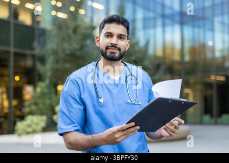 Ein Arzt, wahrscheinlich ein Arzt oder eine Krankenschwester, steht vor einem Gebäude, hält ein Klemmbrett und schaut in die Kamera. Stockfoto