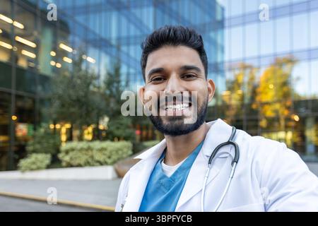 Ein lächelnder Arzt steht vor einem modernen Gebäude, trägt Laborkittel und Stethoskop. Er schaut direkt in die Kamera. Stockfoto