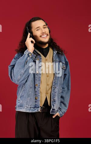 Ein bärtiger junger Mann lächelt und zeigt ein stilvolles Outfit, während er Musik hört. Stockfoto