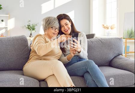 Die alte Mutter und die Erwachsene Tochter sitzen auf dem Sofa und schauen Fotos am Telefon, teilen sich Lachen und schöne Erinnerungen. Alte Mutter und Erwachsene Tochter lieben Stockfoto