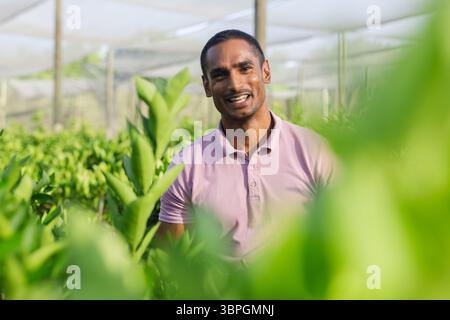 Asiatischer Mann lächelt und trägt rosa Polo im Gewächshaus von Setzlingen unter Schattennetz Stockfoto