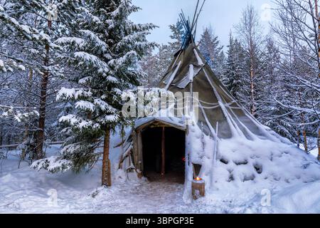 Blick auf ein schneebedecktes Lavvu-Zelt, eingebettet zwischen vereisten Bäumen, ein warmes Licht, das in der Winterdämmerung sanft leuchtet, Rovaniemi, Lappland, Finnland. Stockfoto