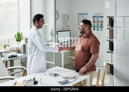 Kaukasier mittleren Alters, der Arzt in der medizinischen Praxis besucht, Hand schüttelt mit jungen erwachsenen südasiatischen männlichen Arzt, diskutiert Diabetes-Management mit medizinischen Geräten auf dem Schreibtisch Stockfoto
