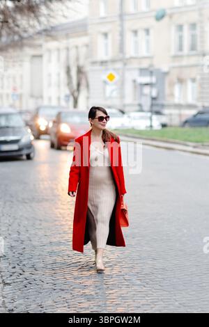 Geschäftsfrau in einem auffälligen roten Mantel und einer stilvollen Sonnenbrille, die selbstbewusst auf einer Kopfsteinpflasterstraße in einem urbanen Stadtbild spaziert Stockfoto