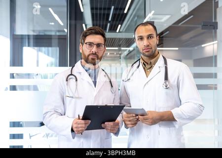 Zwei männliche Ärzte stehen zusammen, einer hält ein Klemmbrett und der andere ein Tablet, in einer modernen medizinischen Umgebung. Stockfoto