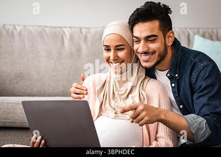 Fröhlich Schwanger Muslimische Frau Und Ihr Mann Mit Laptop An Gemeinsam Zu Hause Stockfoto