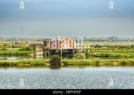 Rustikales Pfahlhaus in einer ruhigen Lagune in Thuy Dien, Hue, Vietnam. Die verwitterte Struktur sitzt auf Holzsäulen und spiegelt sich im ruhigen Wasser. Üppig Stockfoto