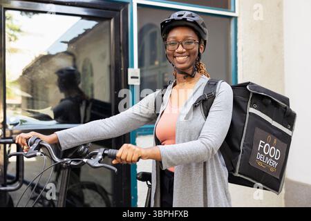 Porträt einer fröhlichen Arbeiterin mit dem Kurierdienst zum Mitnehmen auf dem Fahrrad, mit einem Helm unterwegs und mit ihrem Thermorucksack. Eine Kurierin, die Fast Food- oder Essensbestellungen liefert. Stockfoto