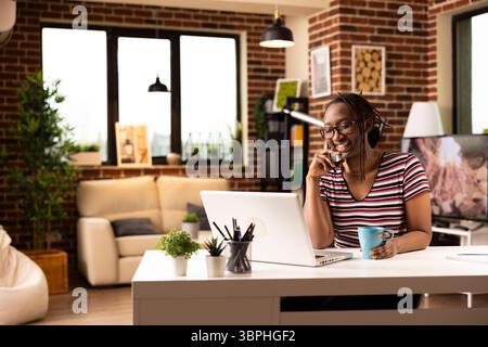 Fröhliche schwarze Frau liest E-Mails auf dem Laptop und teilt während eines Telefonanrufs spannende Neuigkeiten mit Kollegen. Die abgesetzte Managerin sitzt mit einer Kaffeetasse in der Hand am Schreibtisch und spricht während der Arbeit über das Handy. Stockfoto