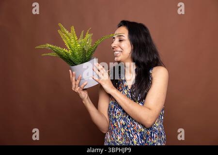 Porträt einer fröhlichen indianerin mit fröhlichem Lächeln, die die Topfpflanze hält und ihre Blätter bewundert. Glückliches weibliches Model steht vor braunem Hintergrund und begeistert mit der Freude an der Gartenarbeit. Stockfoto