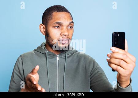 Ein afroamerikanischer Mann, der mit seiner Gruppe von Freunden online telefoniert und über eine Internet-Videokonferenz im Studio plaudert und tratscht. Fröhlicher junger Kerl steht vor blauem Hintergrund. Stockfoto
