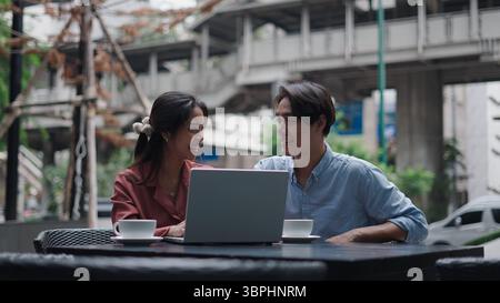 Zwei Geschäftsleute arbeiten gemeinsam an einem Laptop in einem Café im Freien und genießen Kaffee und Unterhaltung Stockfoto