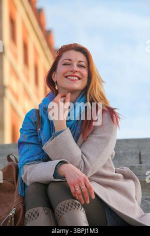 Ingwerhaarige Frau, 30er Jahre, sitzt auf einer Außentreppe, fröhlich und stilvoll in Mantel und Schal. Herbst- oder Frühlingsporträt bei natürlichem Licht. Stockfoto
