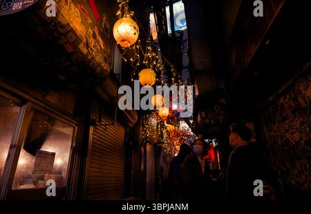 Warme Laternen und Lichterketten beleuchten eine enge Gasse in Tokio, während die Menschen nachts die lebhafte Bar- und Gastronomiebetriebe erkunden Stockfoto