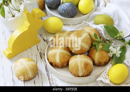 Happy Easter festliches Essenskonzept. Hausgemachte traditionelle heiße Osterbrötchen mit Rosinen und natürlichen bunten ostereiern auf einem rustikalen Tisch Stockfoto
