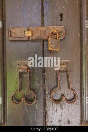 Eine verwitterte Holztür mit rustikalen Metallbeschlägen, einschließlich Verriegelung, Vorhängeschloss und kunstvollen Griffen, die Anzeichen von Alter und Gebrauch zeigen Stockfoto
