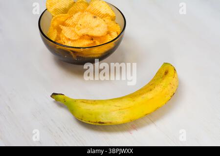 Kartoffelchips in einer Schüssel und eine Banane auf einem weißen Holztisch. Stockfoto