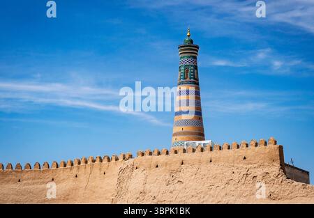 Ein hohes, aufwändig gestaltetes Minarett erhebt sich über einer historischen lehmwand unter einem klaren blauen Himmel. Das Minarett hat bunte Muster und einen goldenen dom Stockfoto
