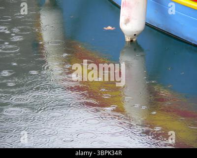 Regentropfen, die auf die Wasseroberfläche fallen, erzeugen Kräuselungen und spiegeln die leuchtenden Farben des Bootsrumpf in einer ruhigen Umgebung im Freien wider Stockfoto