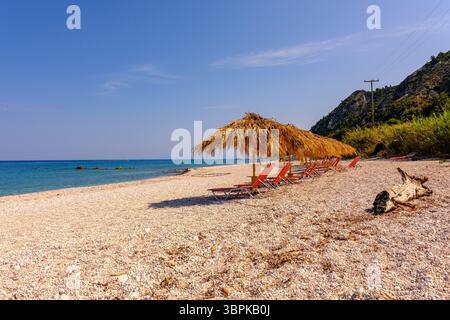 Wunderschöner leerer mediterraner Strand mit Strohschirmen und roten Sonnenliegen, klarem blauem Meer, Kieselstrand und hellem Sommerhimmel. Stockfoto