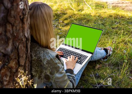 Frau, die im Park am Baum sitzt und einen Laptop benutzt. Leeres grünes Bildschirmmodell, Kopierraum Stockfoto