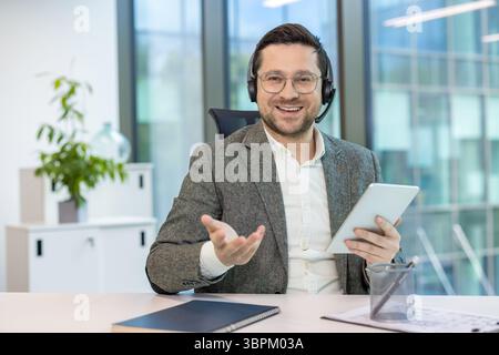 Porträt eines lächelnden erfolgreichen Geschäftsmannes in Headset und Anzug, der an einem Schreibtisch sitzt, ein Tablet in der Hand hält und mit der Kamera spricht. Stockfoto