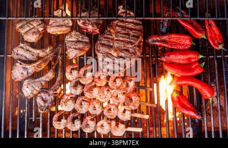 Saftige Steaks und Paprika, die auf einem Grill mit Flammen und aufsteigendem Rauch brutzeln Stockfoto