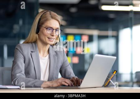 Eine Geschäftsfrau mit Headset arbeitet an einem Laptop in einer modernen Büroumgebung und lächelt beim Tippen. Stockfoto