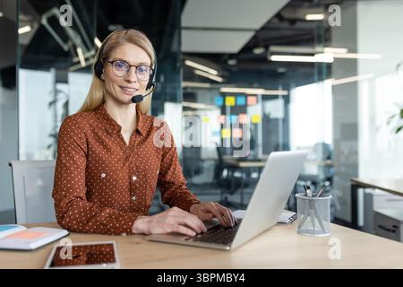 Eine lächelnde blonde Frau, die ein Headset trägt, arbeitet an einem Laptop in einer modernen Büroumgebung. Sie sieht freundlich und hilfsbereit aus. Stockfoto