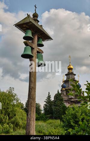 Der traditionelle hölzerne Glockenturm mit grünen Glocken steht vor einer orthodoxen Kirche mit goldenen Kuppeln und Kreuzen, umgeben von Grün unter einer ddr Stockfoto