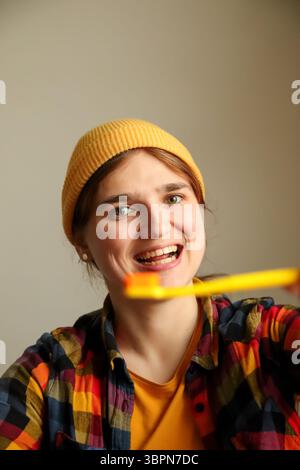 Eine lustige und glückliche Frau mit einer leuchtenden gelben Zahnbürste, die ihre gesunden Zähne in einem breiten Lächeln zeigt, perfekt für eine Dentalanzeige mit Hipster-Vibe. Stockfoto