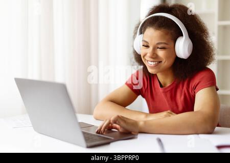 Glückliche junge schwarze Frau im Kopfhörer, die zu Hause online arbeitet oder studiert Stockfoto