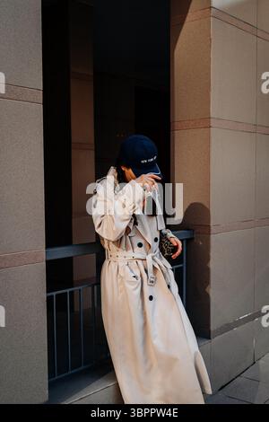 Junge, stilvolle Frau mit trendigem beigefarbenem Regenmantel und blauer Mütze, die durch die Stadt geht, Street-Style, urbane moderne Mode Stockfoto