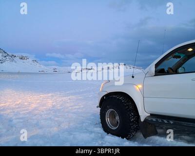 Ein weißer Geländewagen, der während der Dämmerung in einer verschneiten Landschaft geparkt wurde. Die Scheinwerfer des Fahrzeugs beleuchten den Vordergrund und bilden einen Kontrast zu den kühlen Tönen Stockfoto