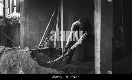 Ein Bauarbeiter, der Sand vor Ort manuell hackt, zeigt Entschlossenheit, körperliche Arbeit und Beharrlichkeit bei der Vorbereitung der Bodenarbeiten. Stockfoto