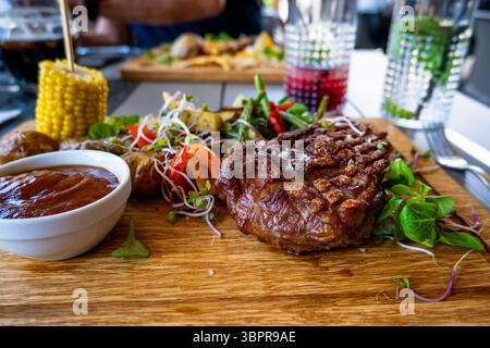Tisch voll mit Essen. Gegrilltes Schweinefleisch, Gemüse, Mais, grünes Blatt, Soße und Getränk auf Holzbrett. Reichhaltiges Mittag- oder Abendessen. Stockfoto