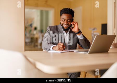 Junger Geschäftsmann lächelt und macht sich Notizen während eines Telefongesprächs, während er an einem Projekt in einem modernen Büro arbeitet Stockfoto
