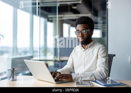 Ein fokussierter Mann mit Brille arbeitet an einem Laptop in einer modernen Büroumgebung und wirkt selbstbewusst und professionell. Stockfoto