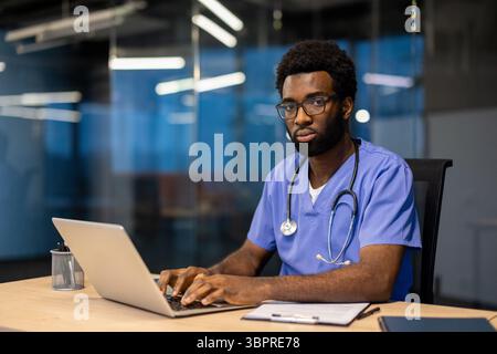 Ein afroamerikanischer Arzt mit Brille und Stethoskop sitzt an einem Schreibtisch in einem Büro und benutzt einen Laptop. Stockfoto