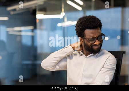 Ein Mann mit einer Brille berührt seinen Hals und scheint im Büro unter Nackenschmerzen zu leiden. Stockfoto