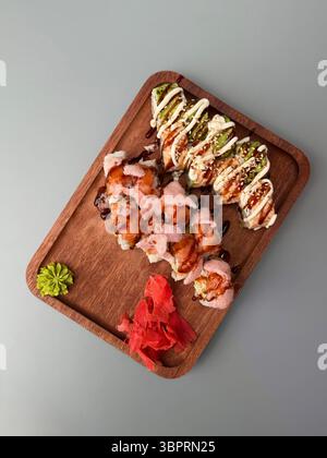Dies ist ein Bild von oben nach unten, das ein Holztablett auf einem hellgrauen Tisch zeigt. Das Tablett enthält zwei Arten von Sushi-Brötchen mit Sauce und Belag Stockfoto