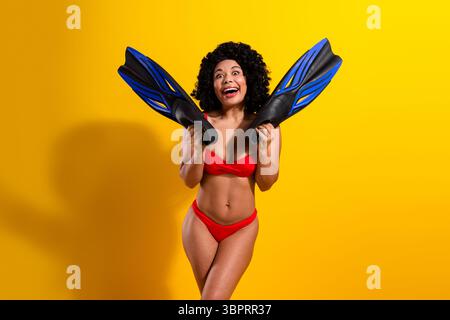 Fröhliche Frau in leuchtend rotem Bikini posiert mit blauen Schwimmflossen vor hellgelbem Hintergrund und bringt Sommerspaß zum Ausdruck Stockfoto