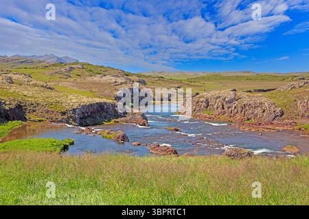 Der klare Fluss fließt durch die felsige Landschaft mit grünen Hügeln in Island unter einem leuchtend blauen Himmel Stockfoto