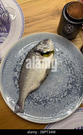 Ganz gebackener Seebarsch auf einem Teller, serviert für ein hausgemachtes Abendessen. Frische Kräuter und Beilagengarnitur schaffen eine warme und einladende Präsentation des Essens. Stockfoto