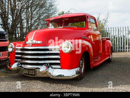 Roter Chevrolet 3100 Pickup-Truck mit Chromakzenten, der auf einer unbefestigten Straße neben einem weißen Streifenzaun und Bäumen geparkt ist Stockfoto