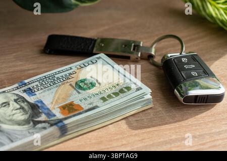 Mit knackigen Hundert-Dollar-Noten neben einem modernen Autoschlüssel auf einer polierten Holzoberfläche, die in sanftes natürliches Licht getaucht ist Stockfoto