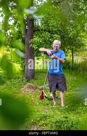 Ein älterer Mann repariert den Rasentrimmer im Garten. Ein älterer Mann mit Schutzbrille führt Wartungsarbeiten an einem Rasentrimmer in einem üppigen, grünen Hinterhof durch Stockfoto