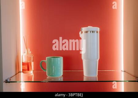 Grüner Keramikbecher, minimalistischer weißer Becher und Reed-Diffusor zur Dekoration des Glasregals vor der beleuchteten roten Wand. Stockfoto